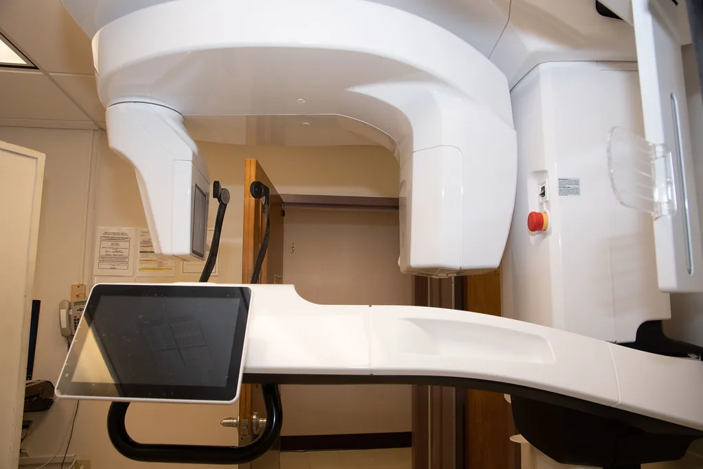 An upright cone beam CT scanner in a clinical imaging room, with its rotating gantry and touchscreen console